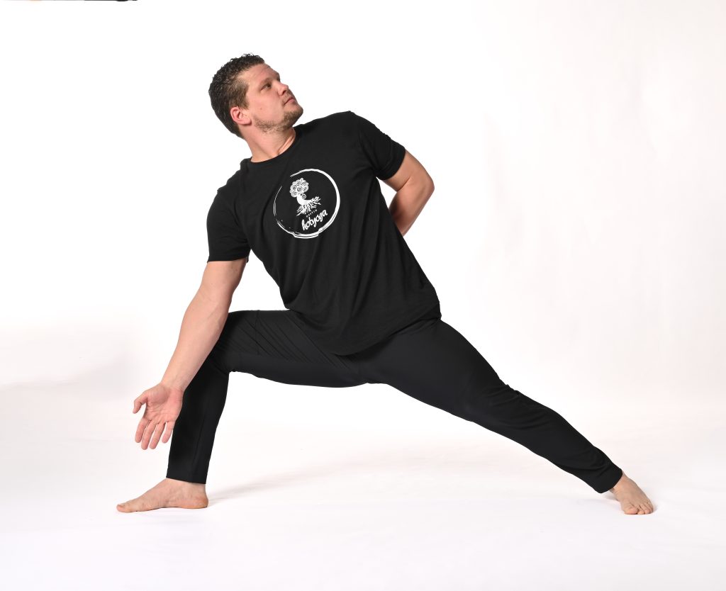 A man in a black shirt and pants, doing a yoga stretch. He is a wearing a Destin Hot Yoga boutique shirt.